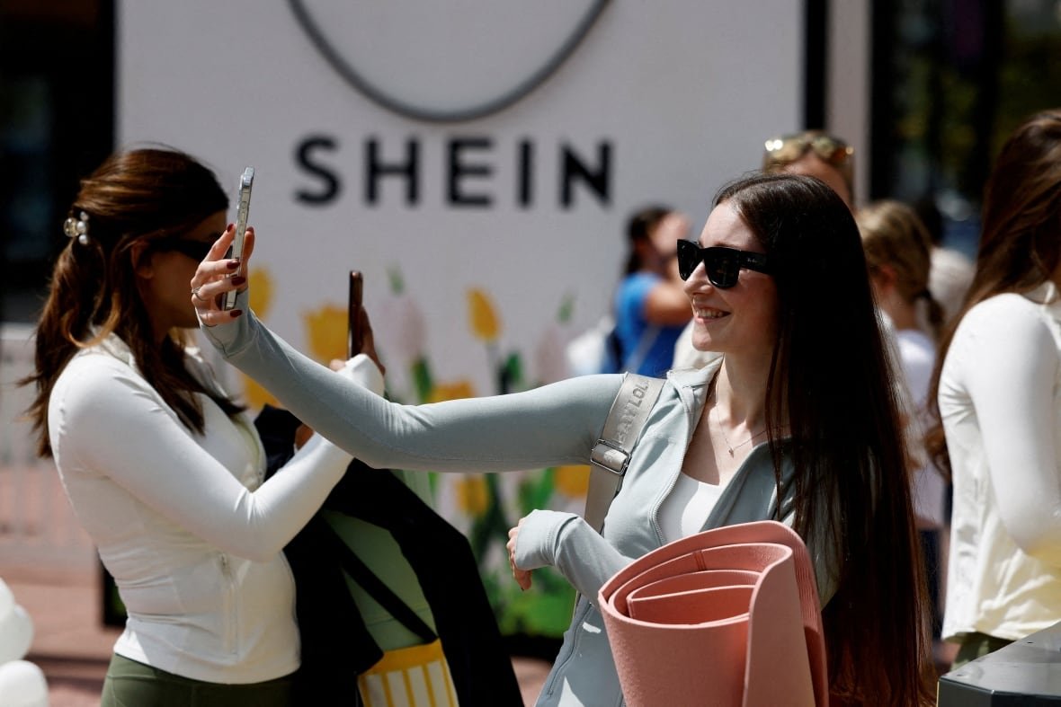 shein-ipo-labour Women stand in front of a sign reading 'Shein' with their phones out, taking pictures.
