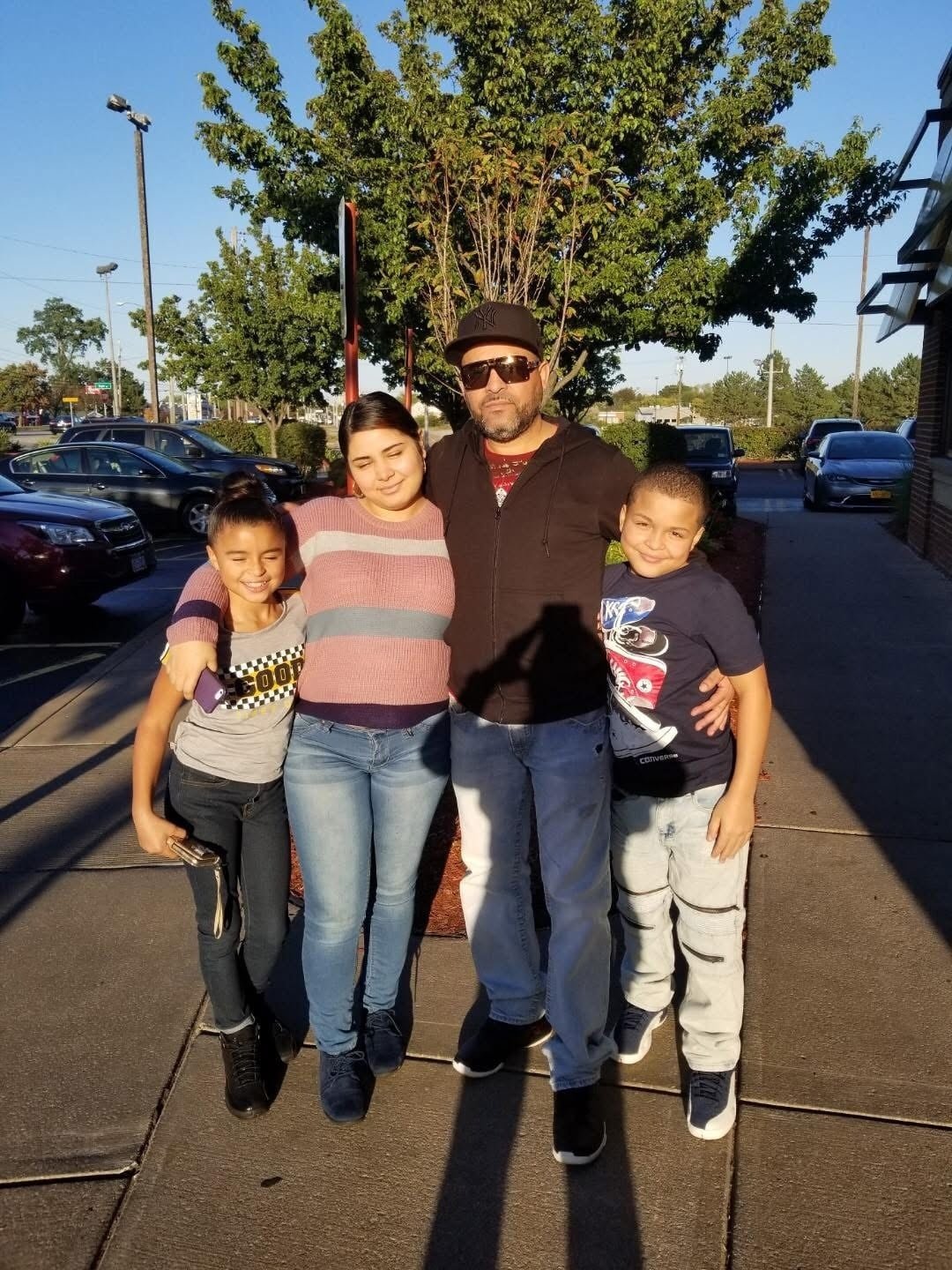 A bearded man in sunglasses and a cap has his arms around three children, which appear to be two boys and a girl.