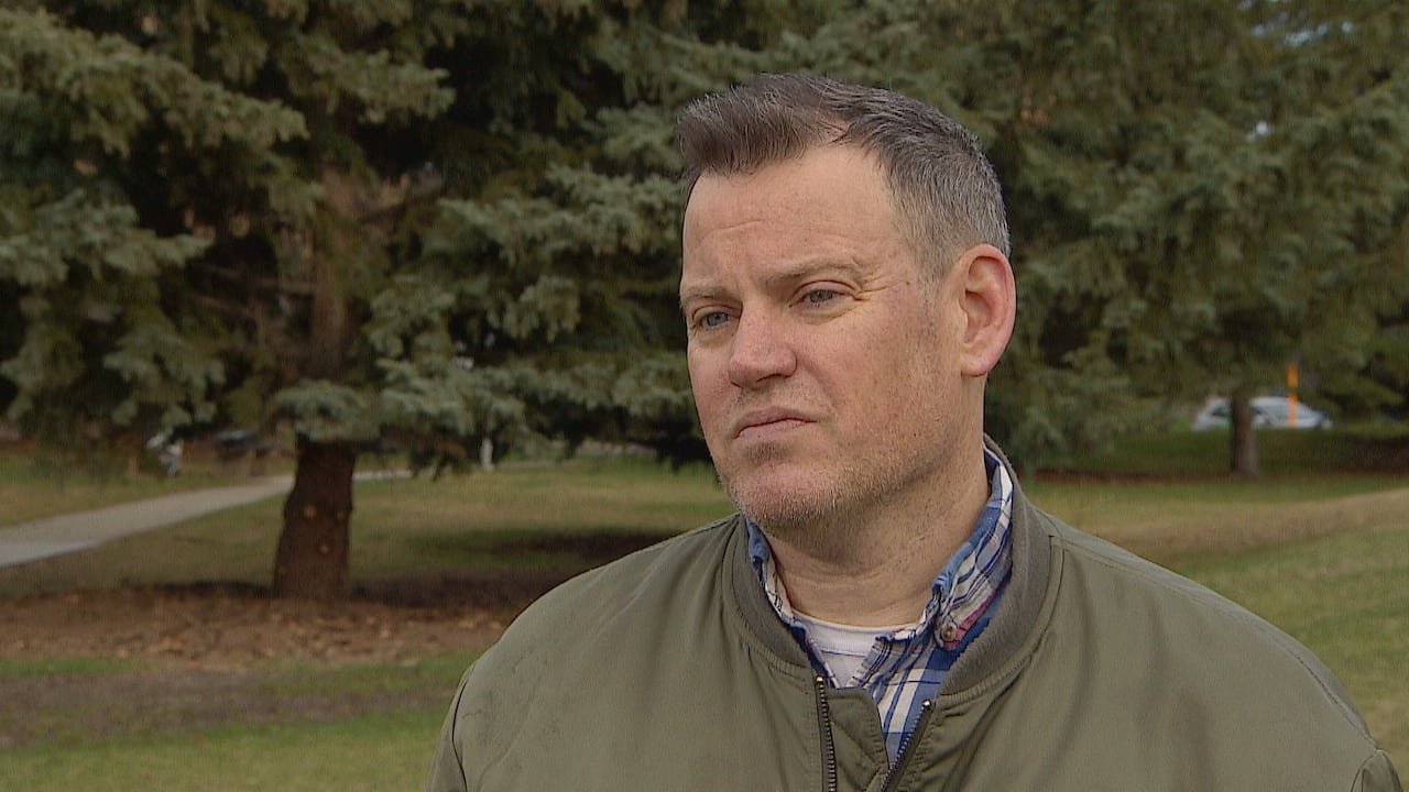 A man wearing a green jacket stands against a backdrop of trees.
