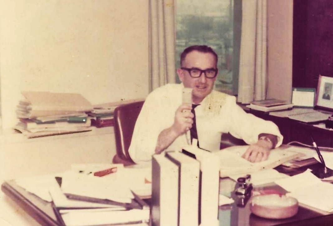 A vintage photograph of a man seated at a desk holding a telephone