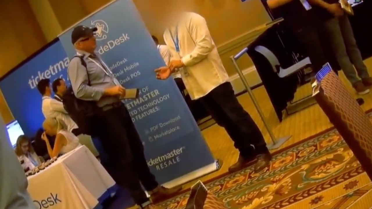 An undercover journalist speaks to a man with a blurred face on the convention floor of a ticket industry convention.