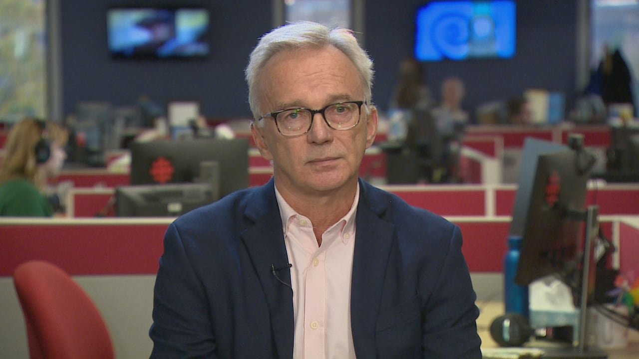 default-234 A man in glasses wears a blue blazer and light pink shirt as he sits on a TV news set.