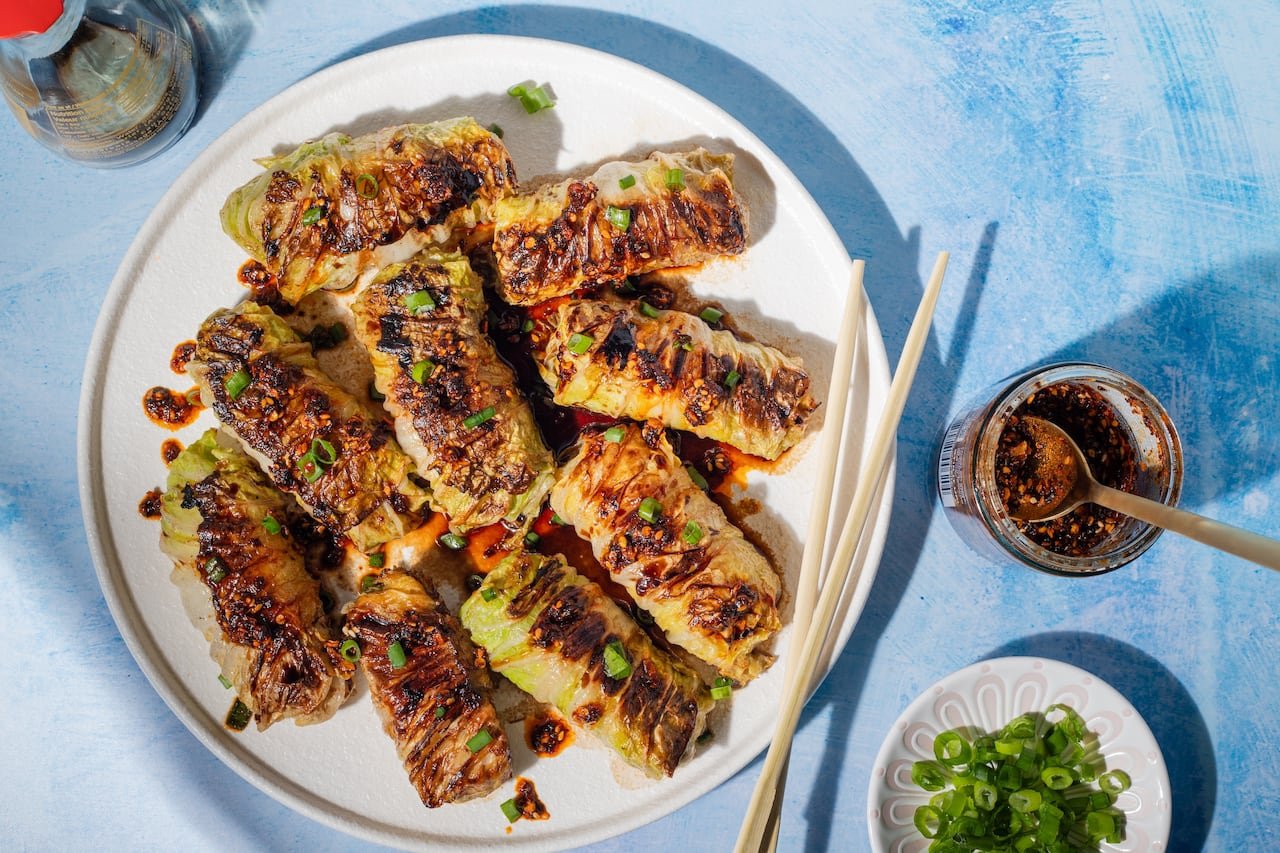 default-689 Top-down view of a white platter filled with dumplings wrapped in cabbage leaves, drizzled with soy sauce and garnished with green onions, placed on a blue surface.