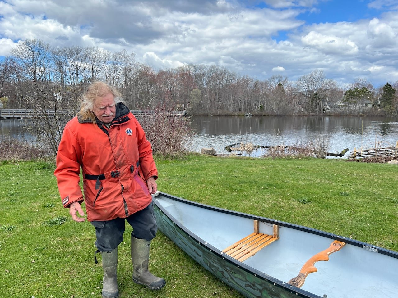 An older man with white hair and a bright orange jacket beside a small boat on grass.