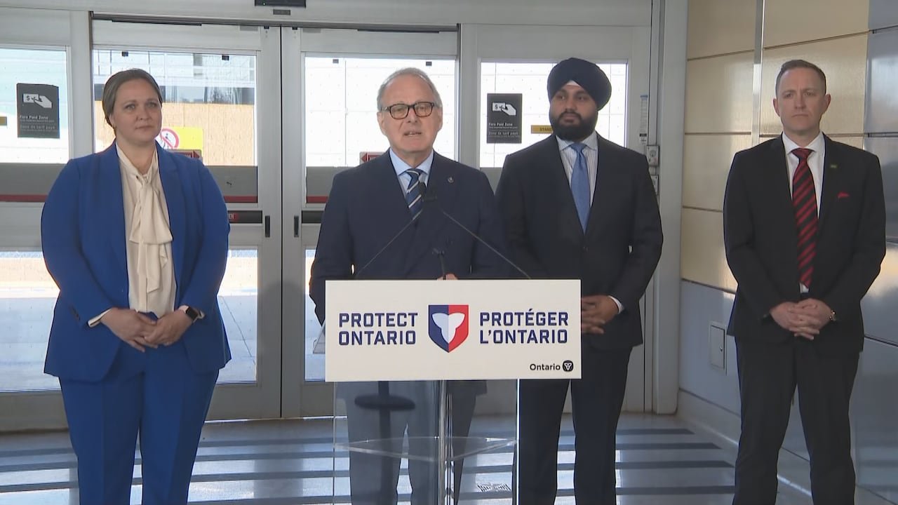 A middle-aged white man in a suit and tie speaks at a lectern set up in a Toronto subway station. A woman and two men in business attire stand behind him