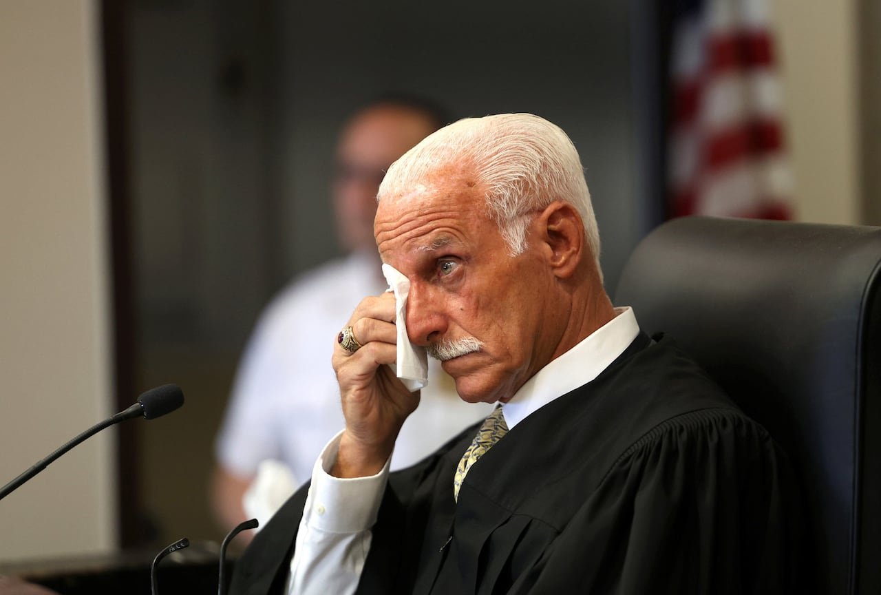 A balding man with white hair and white mustache in judicial robe wipes his eye while seated.