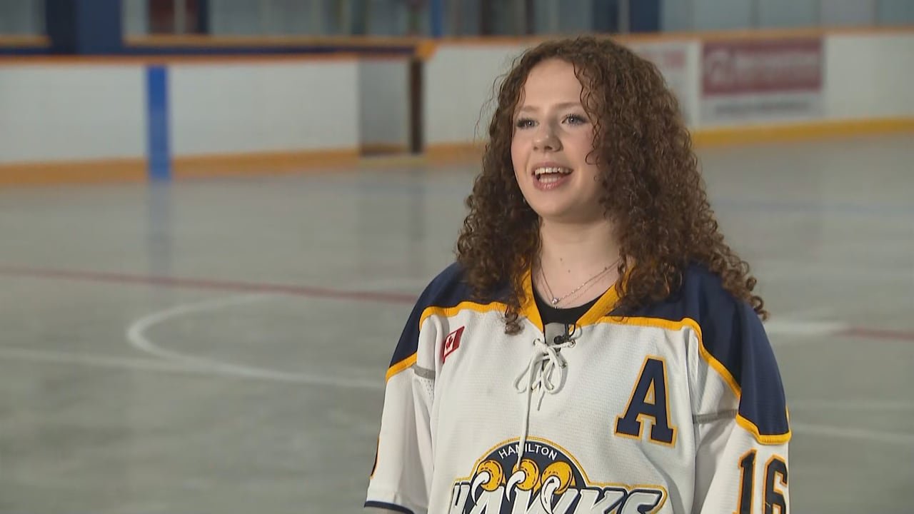 A teenage girl with curly hair stands in an empty arena wearing a Hawks #16 jersey.