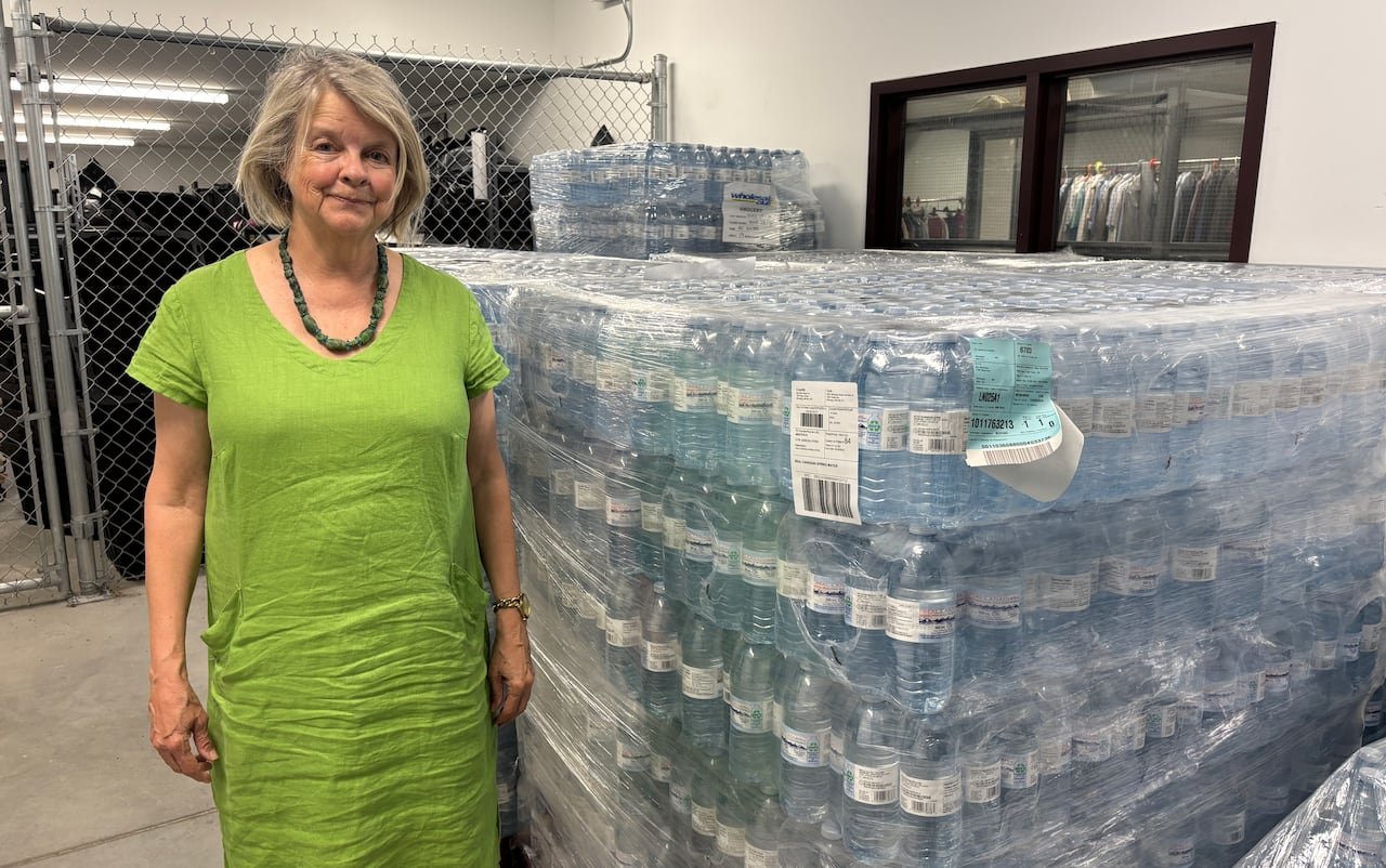 default-974 A woman in a long green dress smiles and stands next to large pallets full of plastic water bottles.