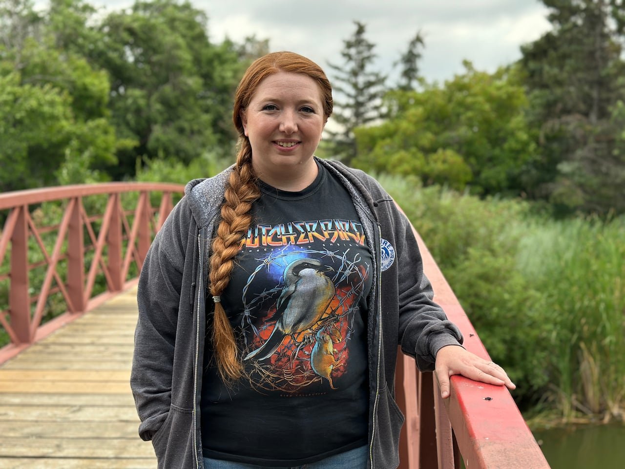 A woman with a long braid stands on a small bridge crossing a creek, smiling at the camera. She is wearing a shirt with a graphic depicting the "butcher bird," or loggerhead shrike.