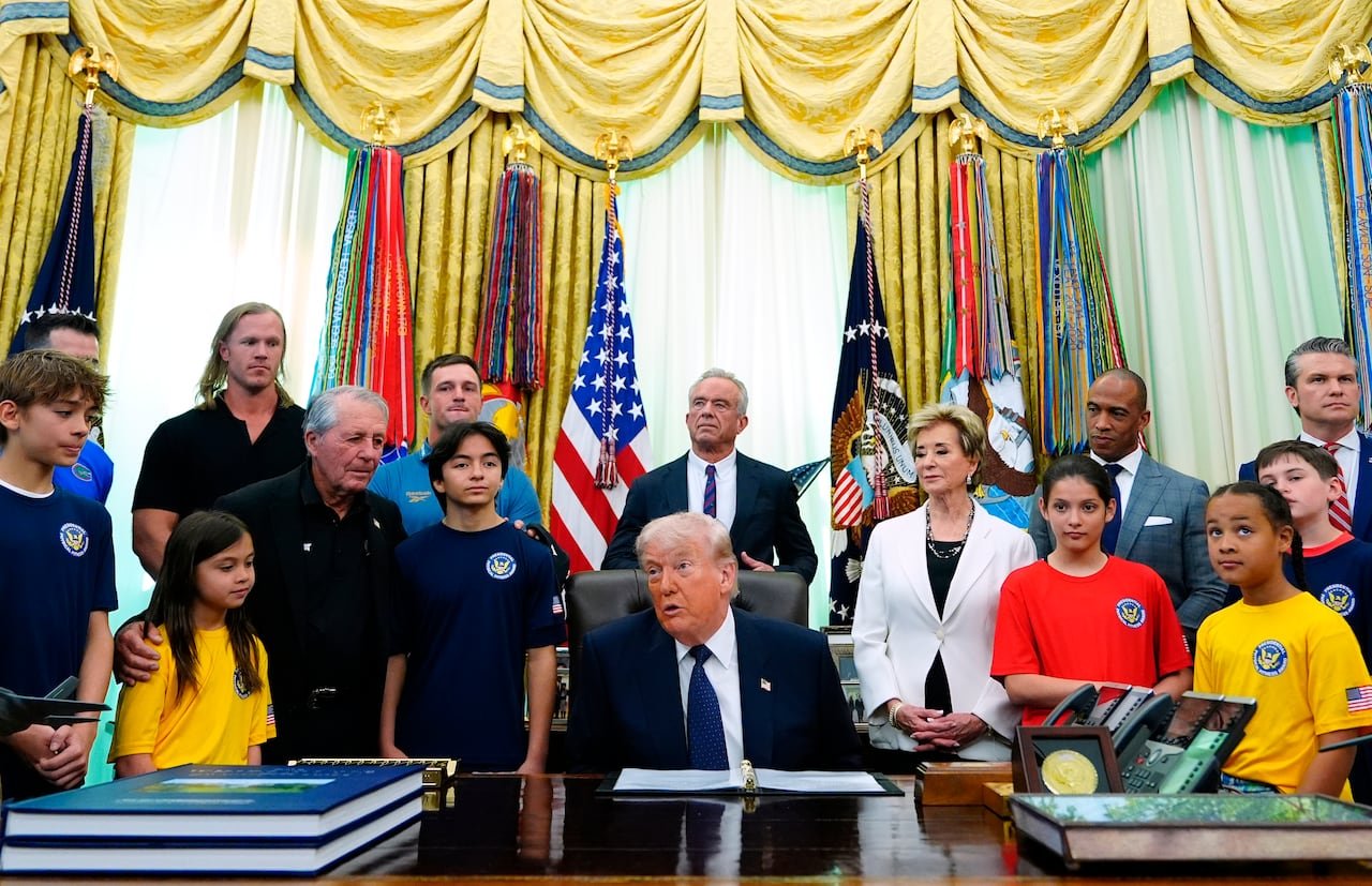 default-394 Donald Trump is sitting at his desk in the Oval Office while surrounded by some children and a few members of his cabinet.