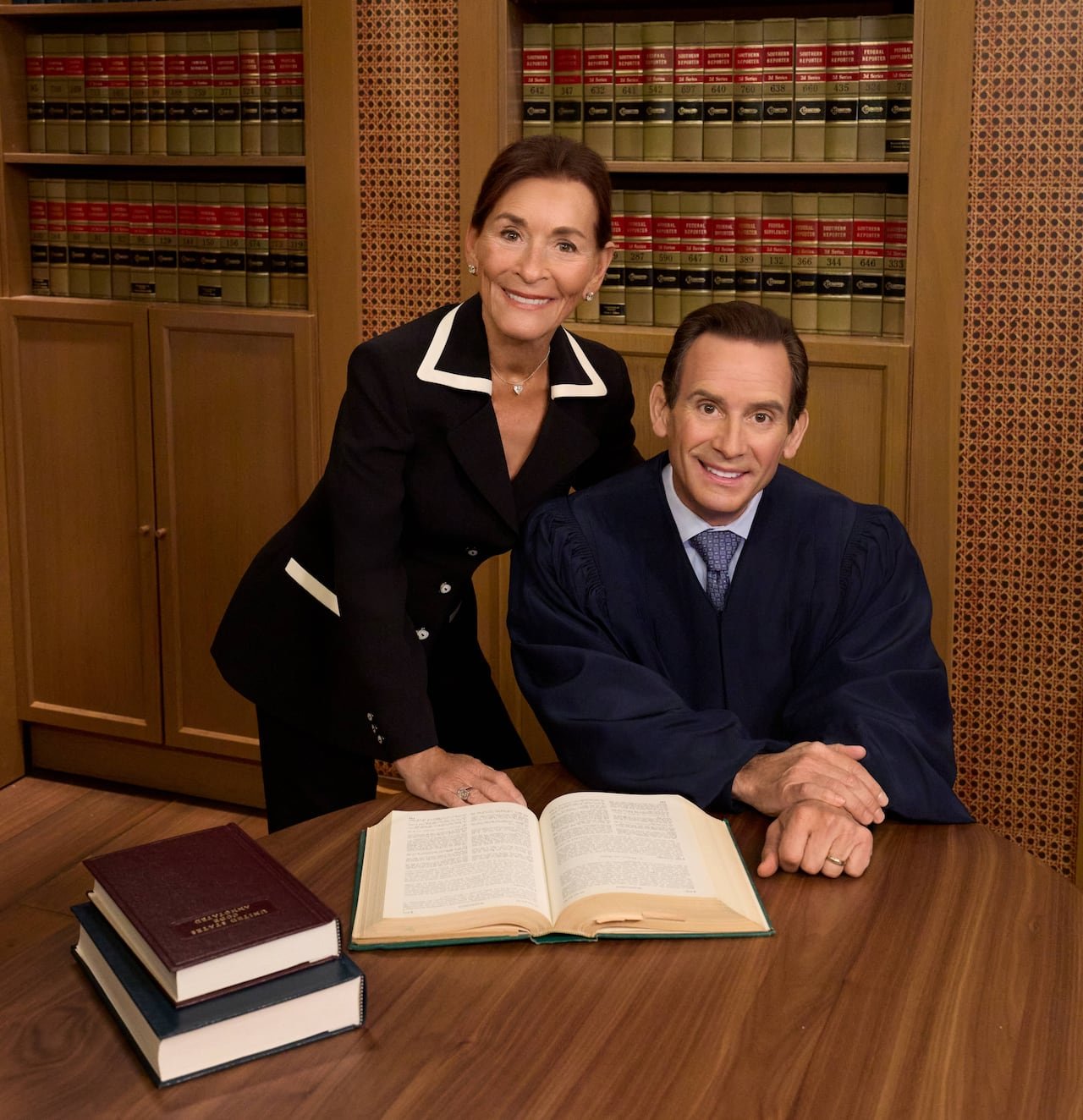 default-446 A woman in a black suit jacket leans over a man in a judge's robe as he's seated at a desk, with a book open in front of him. Both are smiling directly at the camera.