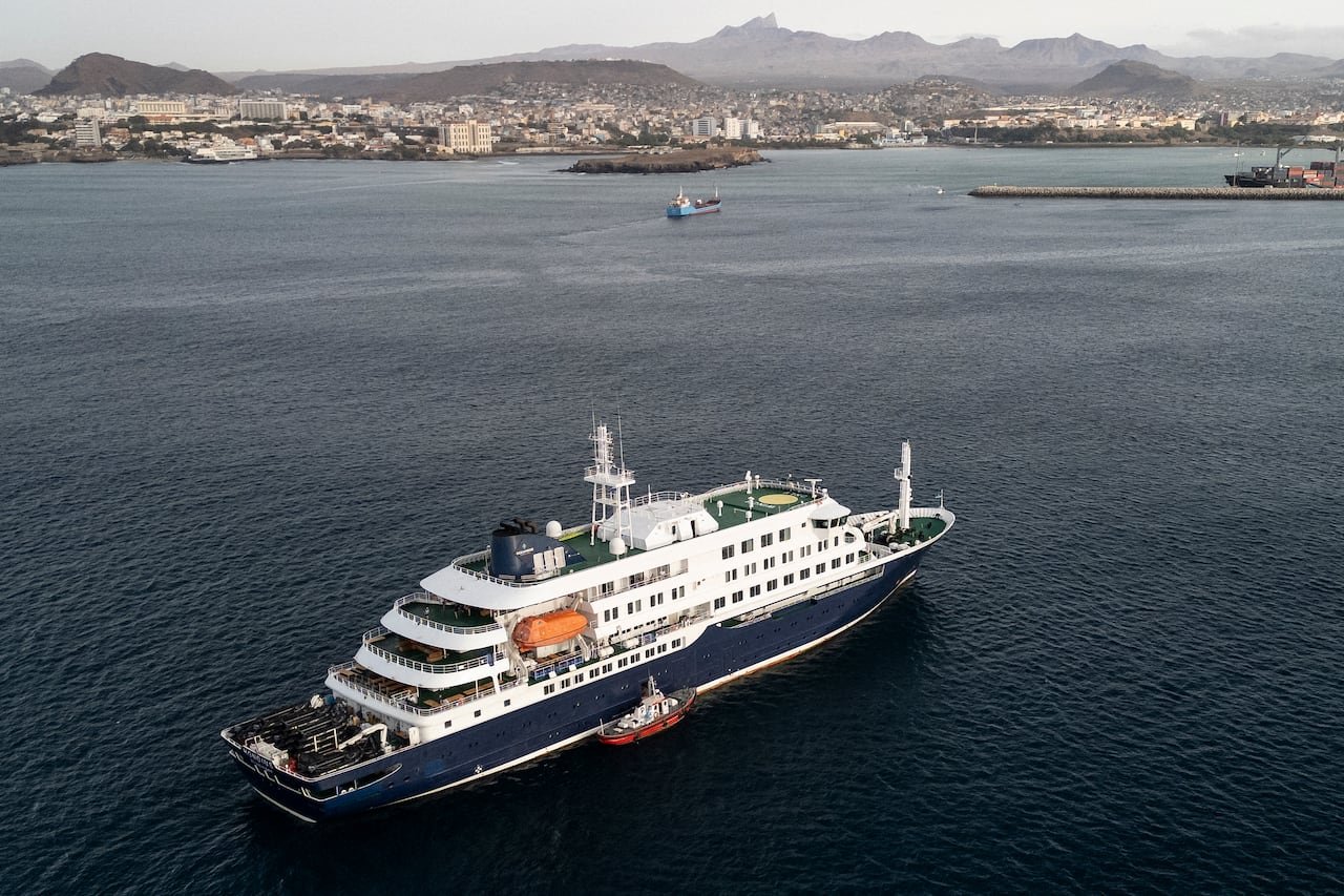 An aerial shot of small boat beside a white and blue cruise ship anchored on the blue water offshore, with buildings lining on a mountainous island in the background.