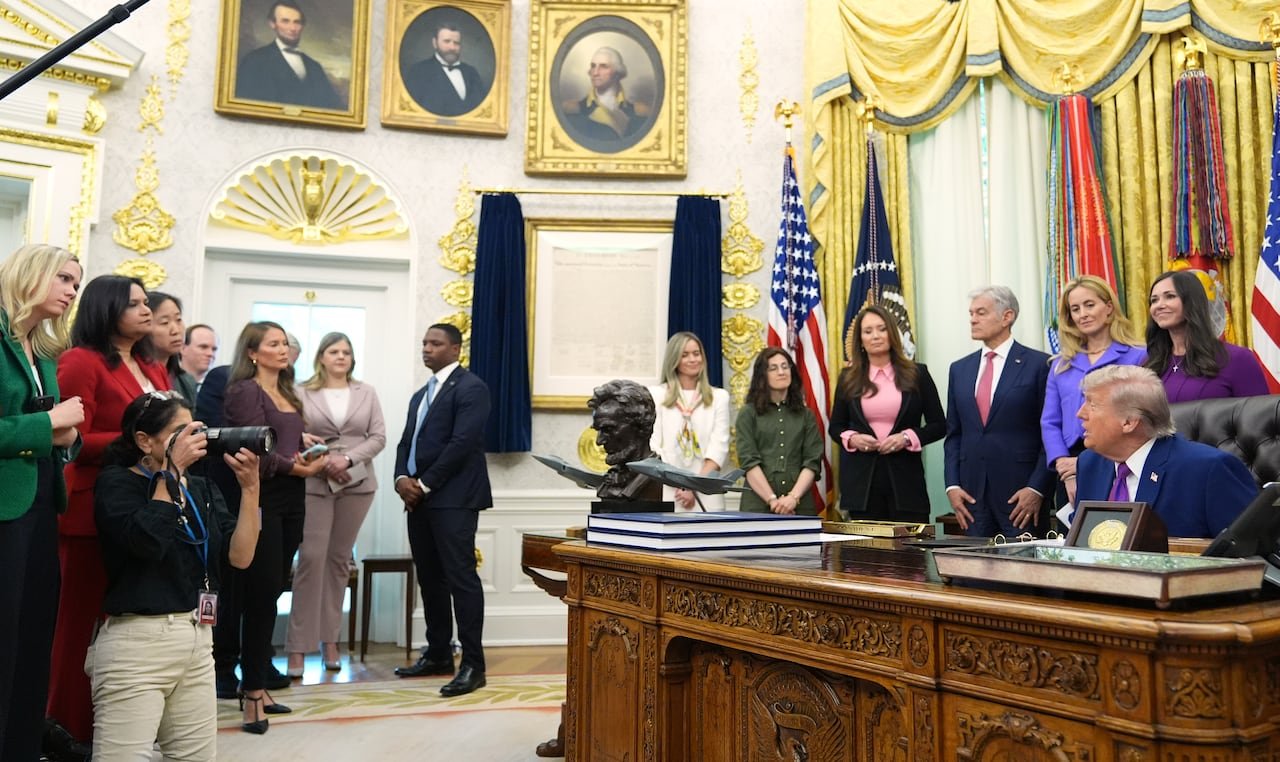 default-257 A wide-angle photo in the Oval Office shows Donald Trump sitting at his desk, looking across toward a group of reporters, with gold-framed paintings of presidents on the wall behind.