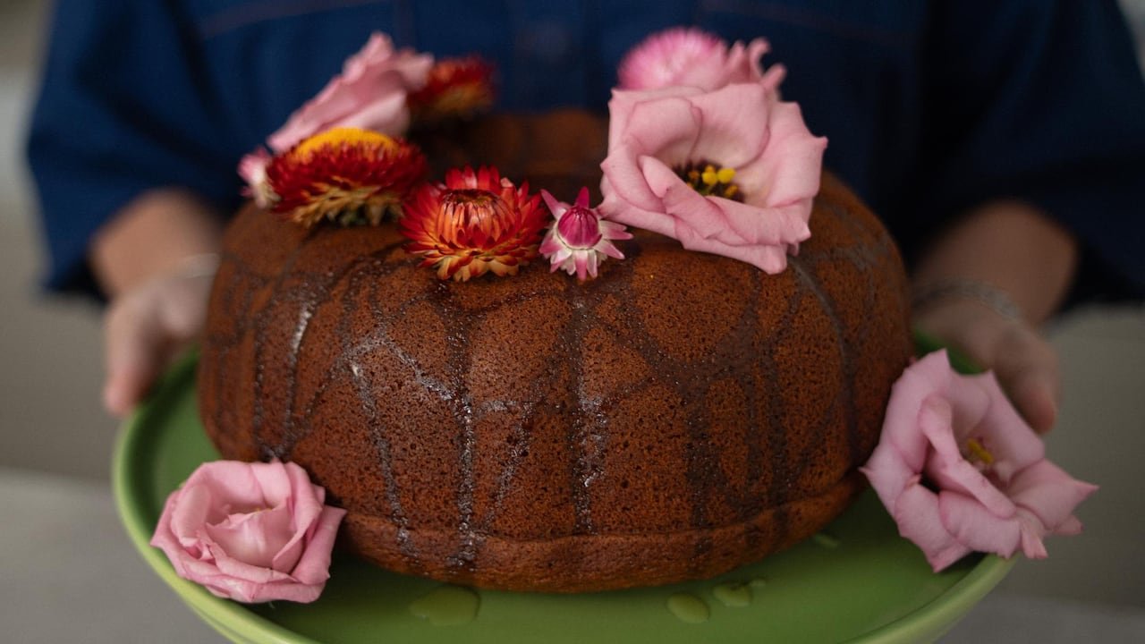 default-498 Closeup of a honey-drizzled bundt cake adorned with pink flowers on a green cake plate held by hands wearing a navy blue shirt.