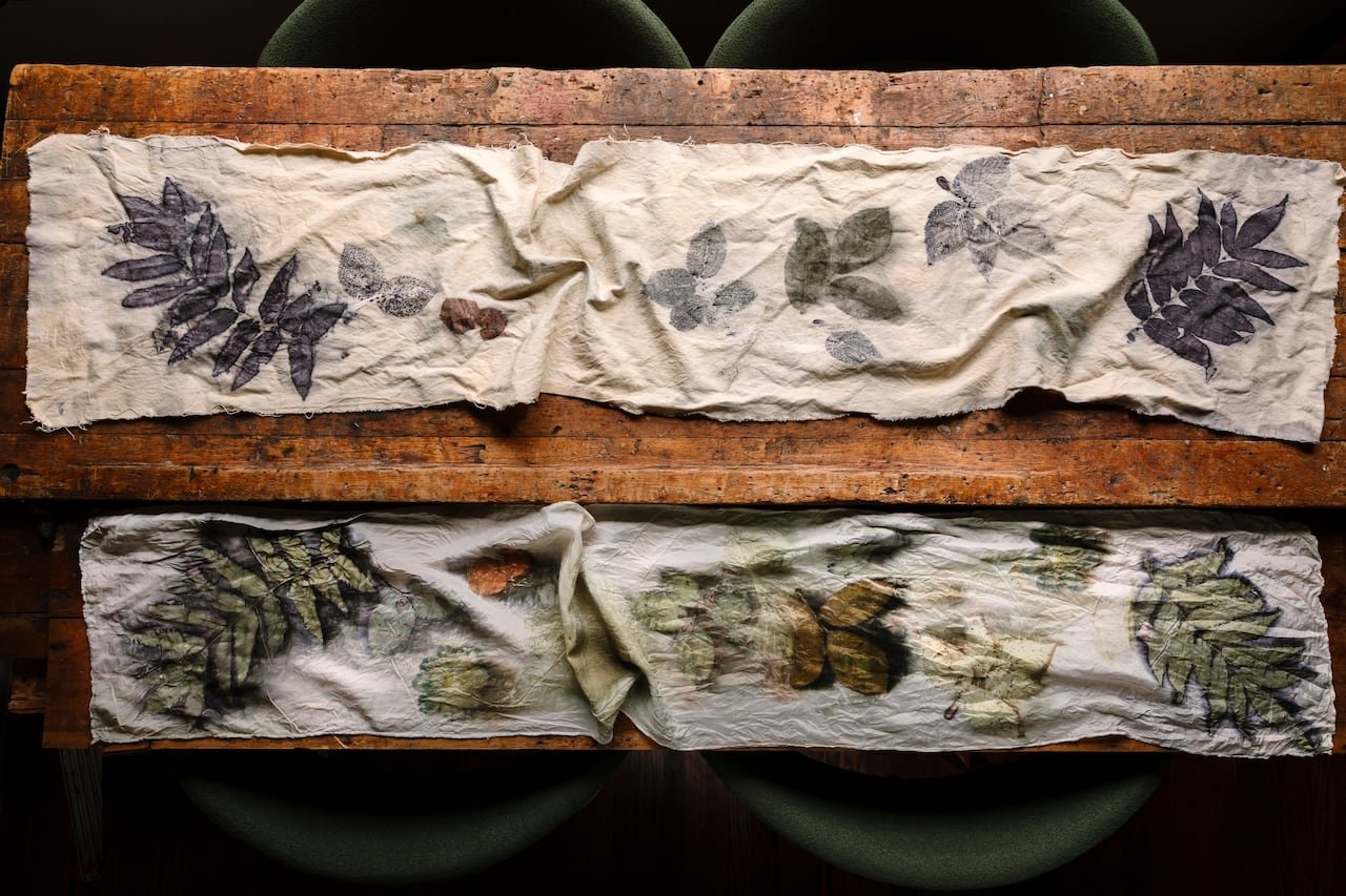 A wooden table with 2 table runners on it. They both have dyed prints of foliage.